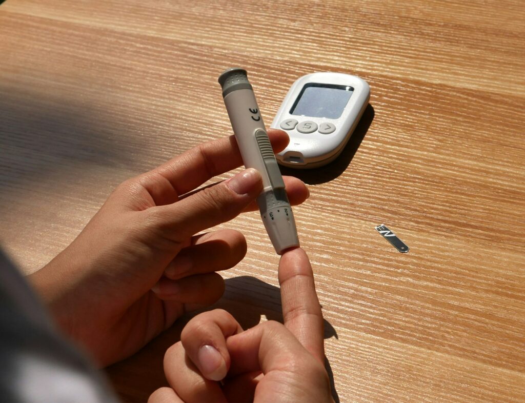 A pregnant woman’s hands using a blood glucose finger prick test, with a glucose meter and test strip on a wooden table.