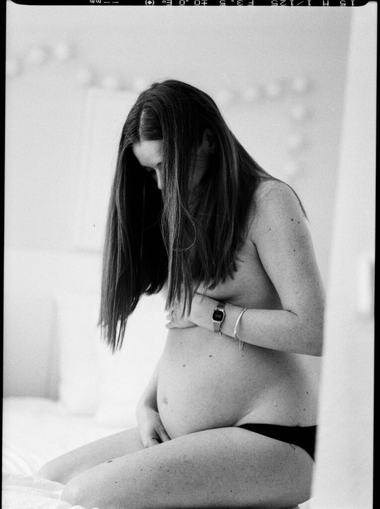 Black and white photograph of a pregnant woman sitting on a bed, cradling her belly with both hands. Her long hair falls over her face as she looks down, appearing calm and introspective. She wears a smartwatch and bracelets on her wrist, and the soft, blurred background creates a peaceful, intimate atmosphere.