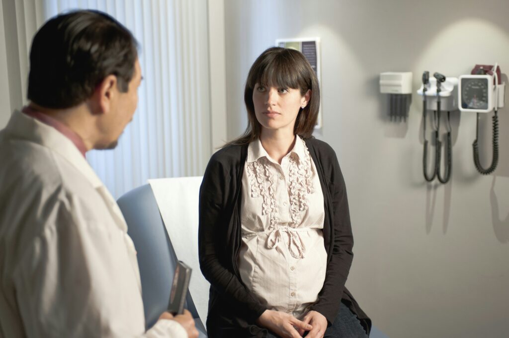 Pregnant woman sitting in a medical exam room speaking with a doctor, looking serious and slightly tense during a prenatal appointment.